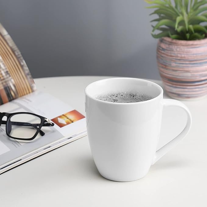 A white ceramic mug filled with a hot beverage, placed on a white table next to eyeglasses, a magazine, and a potted plant. no-save