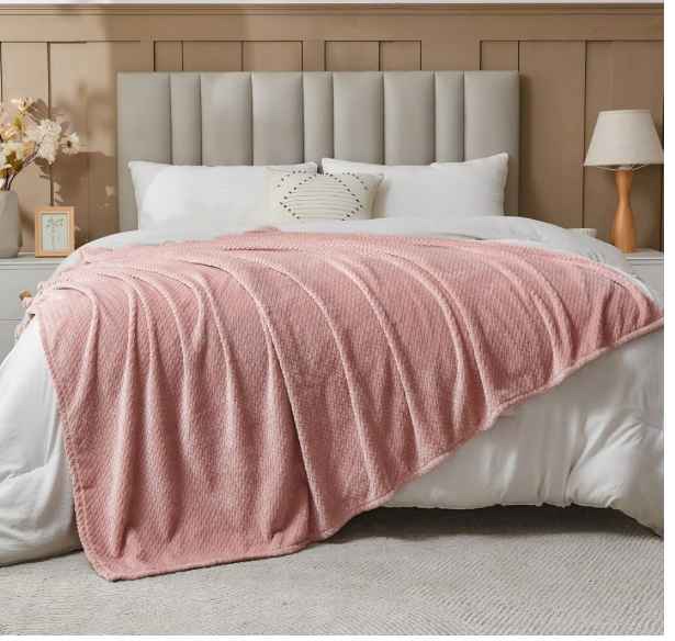 A beautifully styled bedroom featuring a plush dusty pink throw blanket draped over a white bed. The space is complemented by soft neutral tones, an upholstered headboard, warm lighting from a wooden bedside lamp, and delicate floral accents for a cozy and elegant atmosphere. no-save