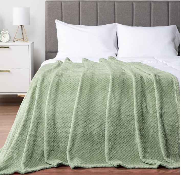 A cozy sage green knitted blanket draped over a neatly made bed with white pillows and bedding. The bedroom features a modern, neutral-toned headboard and a minimalist nightstand with a lamp and decorative items, creating a warm and inviting atmosphere. no-save