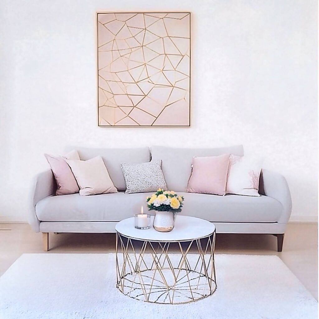 An elegant living room with a minimalist gray sofa, accented with blush and gold pillows, a modern geometric coffee table, and a floral centerpiece under soft ambient light.