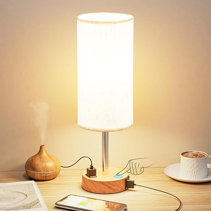 A modern bedside lamp with a wooden base, touch control, and USB charging ports, emitting a warm glow. A phone is charging nearby, alongside a steaming cup of coffee and an essential oil diffuser, creating a cozy and functional nightstand setup. no-save