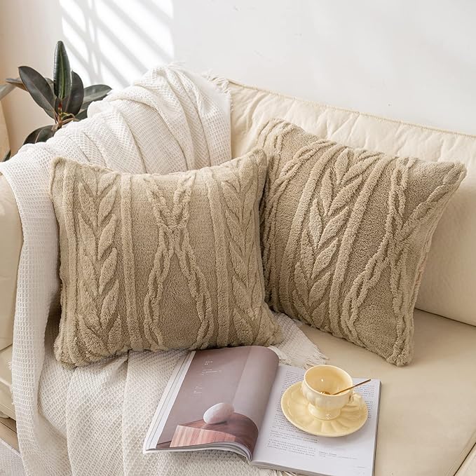 Two cozy tan throw pillows with a textured leaf pattern resting on a cream-colored sofa. A white knitted blanket, an open magazine, and a delicate yellow teacup add to the inviting and serene ambiance of the space. no-save