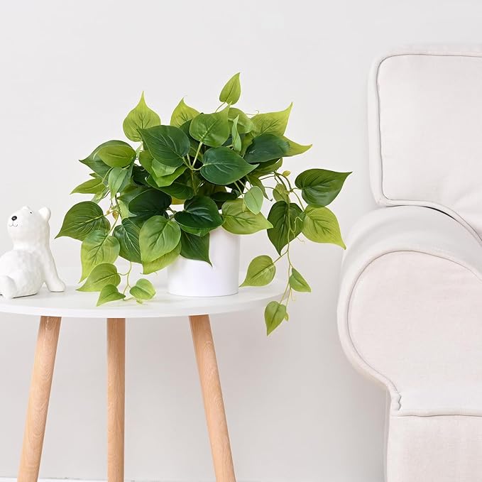 Artificial green pothos plant in a white pot, placed on a modern wooden side table next to a cozy white armchair, adding a touch of freshness to the space. no-save